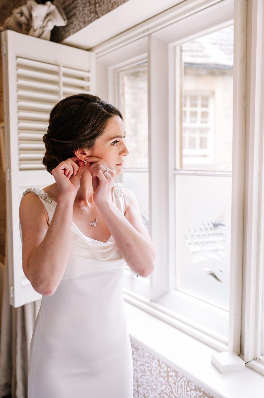 SUMMER WEDDING AT THE SWAN HOTEL BIBURY 15 Close-up of a bride finishing her bridal look by putting on earrings for her summer wedding at the Swan Hotel Bibury.