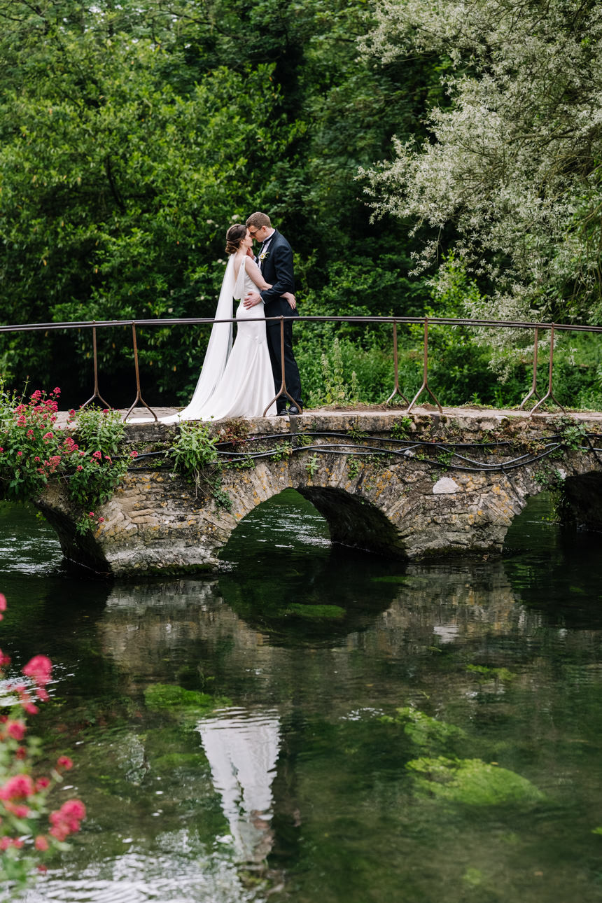 SUMMER WEDDING AT THE SWAN HOTEL BIBURY 33 Iconic portrait of the bride and groom standing on a traditional stone footbridge during their summer wedding at the Swan Hotel Bibury.