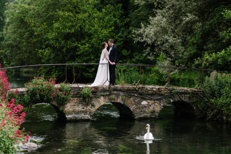Iconic portrait of the bride and groom standing on a traditional stone footbridge during their summer wedding at the Swan Hotel Bibury.