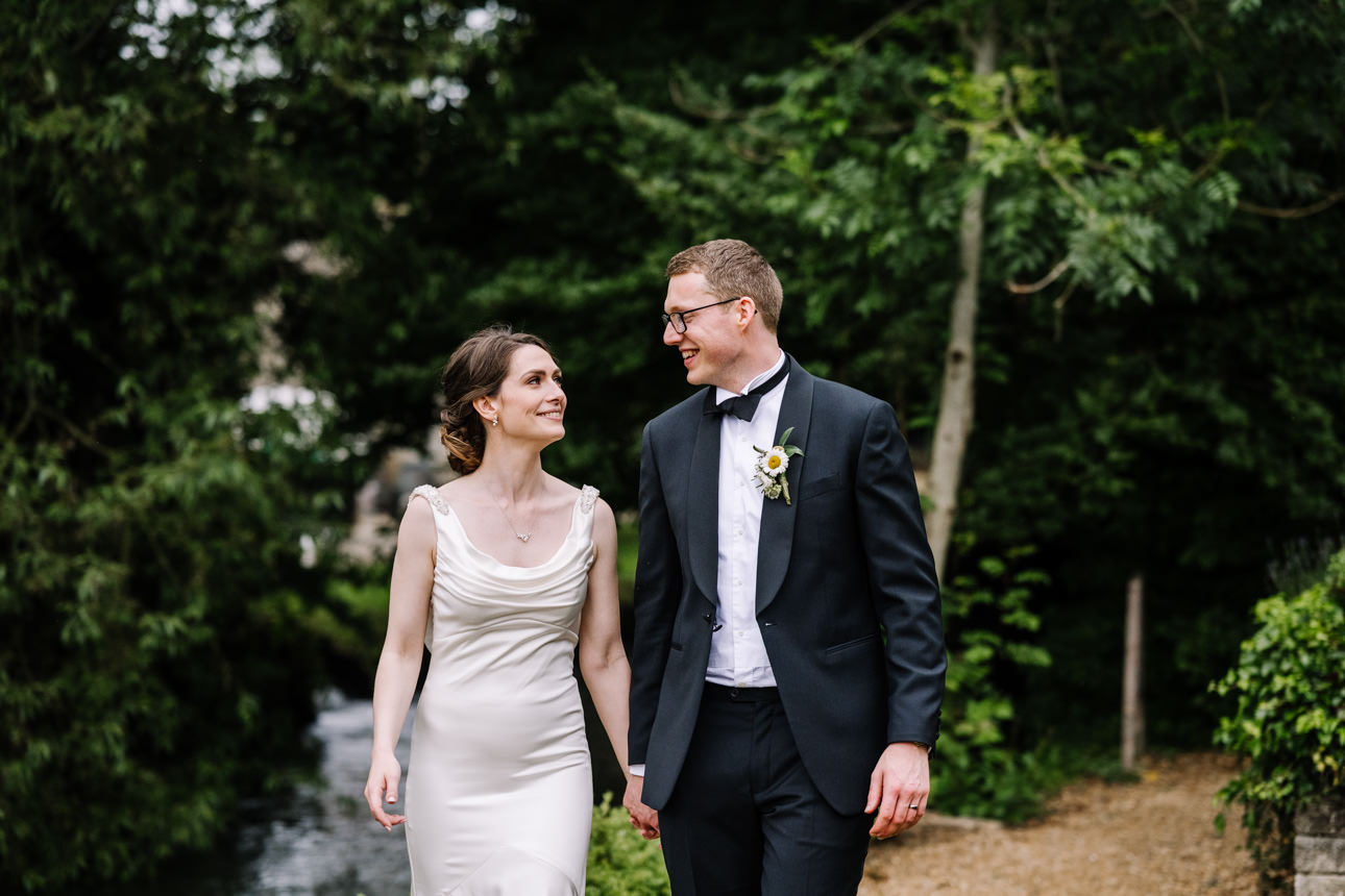 SUMMER WEDDING AT THE SWAN HOTEL BIBURY 51 Bride and groom walking along the lush green river bank at their summer wedding at the Swan Hotel Bibury.