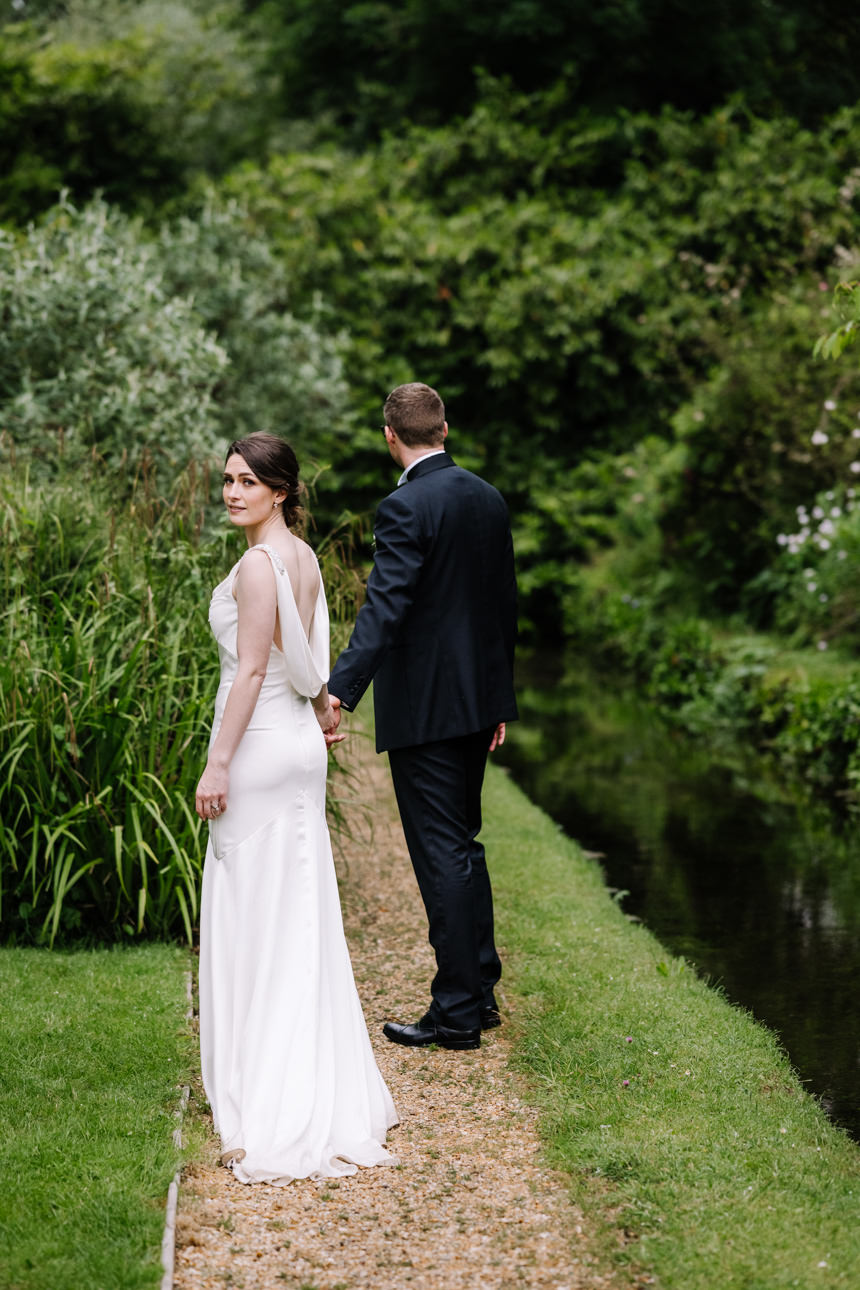 SUMMER WEDDING AT THE SWAN HOTEL BIBURY 50 Bride and groom walking along the lush green river bank at their summer wedding at the Swan Hotel Bibury.
