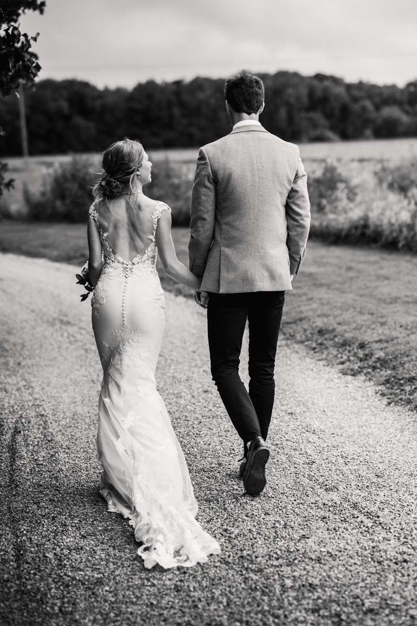 LAPSTONE BARN WEDDING PHOTOGRAPHER 50 Rear-view black and white photograph of a bride and groom walking hand-in-hand down a lane at Lapstone Barn.