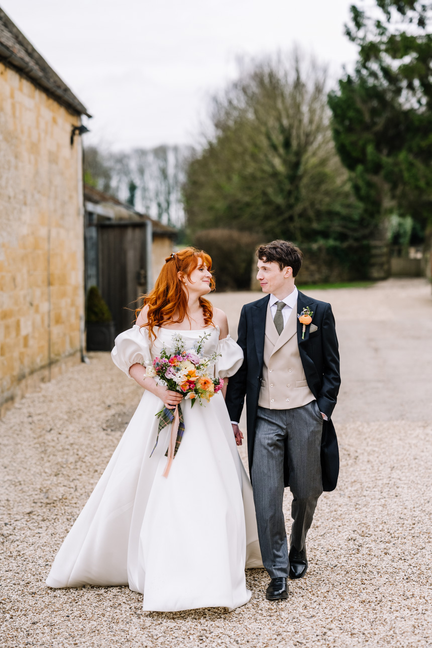 LAPSTONE BARN WEDDING PHOTOGRAPHER 56 A bride with a long dark train walking hand-in-hand with her groom near the stone buildings of Lapstone Barn.