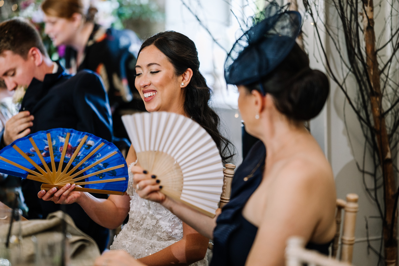 LAPSTONE BARN WEDDING PHOTOGRAPHER 79 Female wedding guests using hand fans during a warm summer wedding reception inside Lapstone Barn.