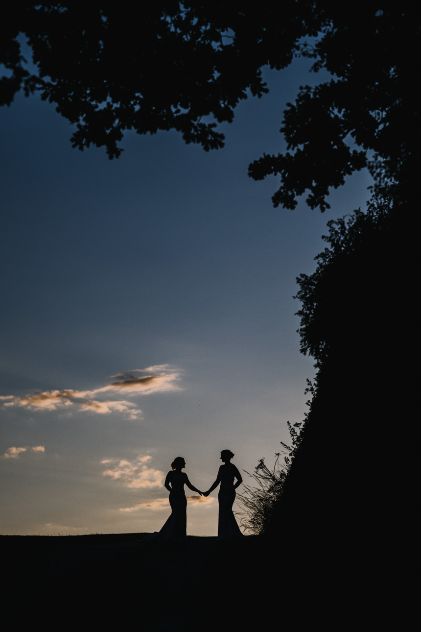KINGSCOTE BARN WEDDING PHOTOGRAPHER 65 Silhouette wedding photography of two brides holding hands against a twilight sky, captured by Kingscote Barn wedding photographer Tanli Joy Weddings.