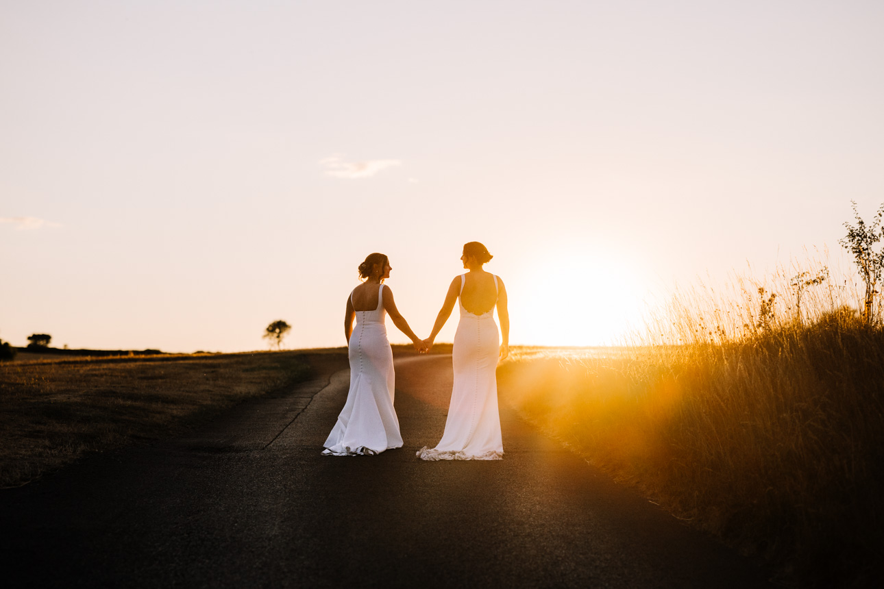 KINGSCOTE BARN WEDDING PHOTOGRAPHER 66 Two brides in elegant white wedding dresses walking hand-in-hand down a path during a golden hour sunset at Kingscote Barn.