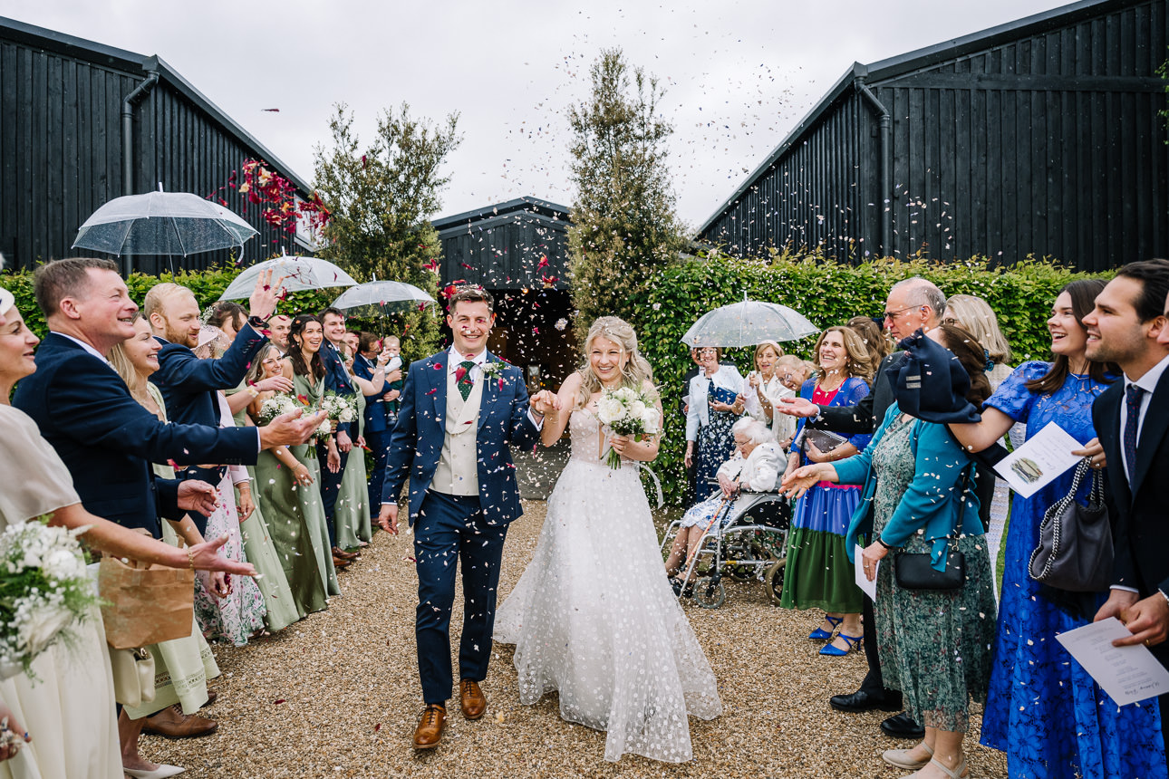 FLORA AND WILLIAM PRIMROSE HILL FARM SPRING WEDDING 35 Newly married couple walking through colourful confetti surrounded by cheering wedding guests