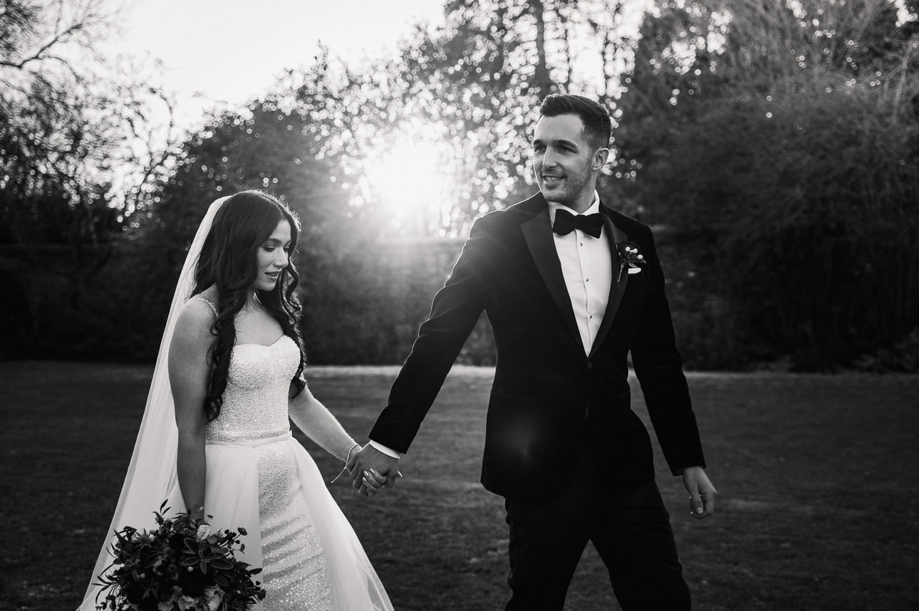 NATURAL WEDDING PHOTOGRAPHY 42 A dramatic black and white photograph of a bride and groom holding hands and walking together down a rustic path at sunset.