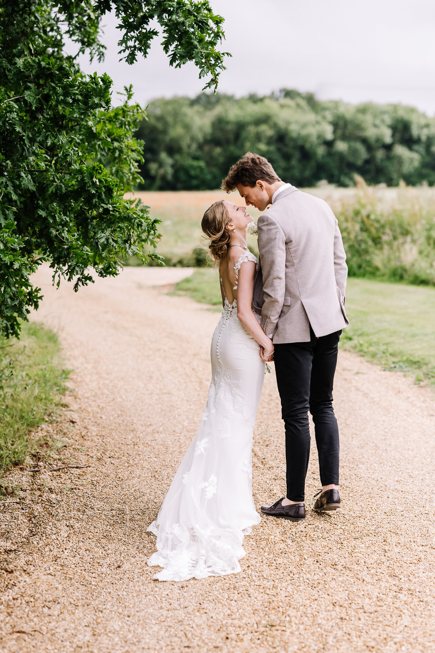 NATURAL WEDDING PHOTOGRAPHY 43 A bride and groom walking together down a curved gravel pathway bordered by lush green fields on their wedding day.