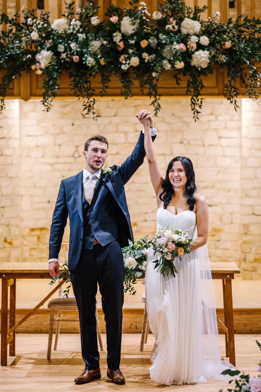 GREAT TYTHE BARN WEDDING PHOTOGRAPHER 25 Newlyweds Greg and Chloe cheering with raised hands as they walk down the aisle together inside the main hall of The Great Tythe Barn.