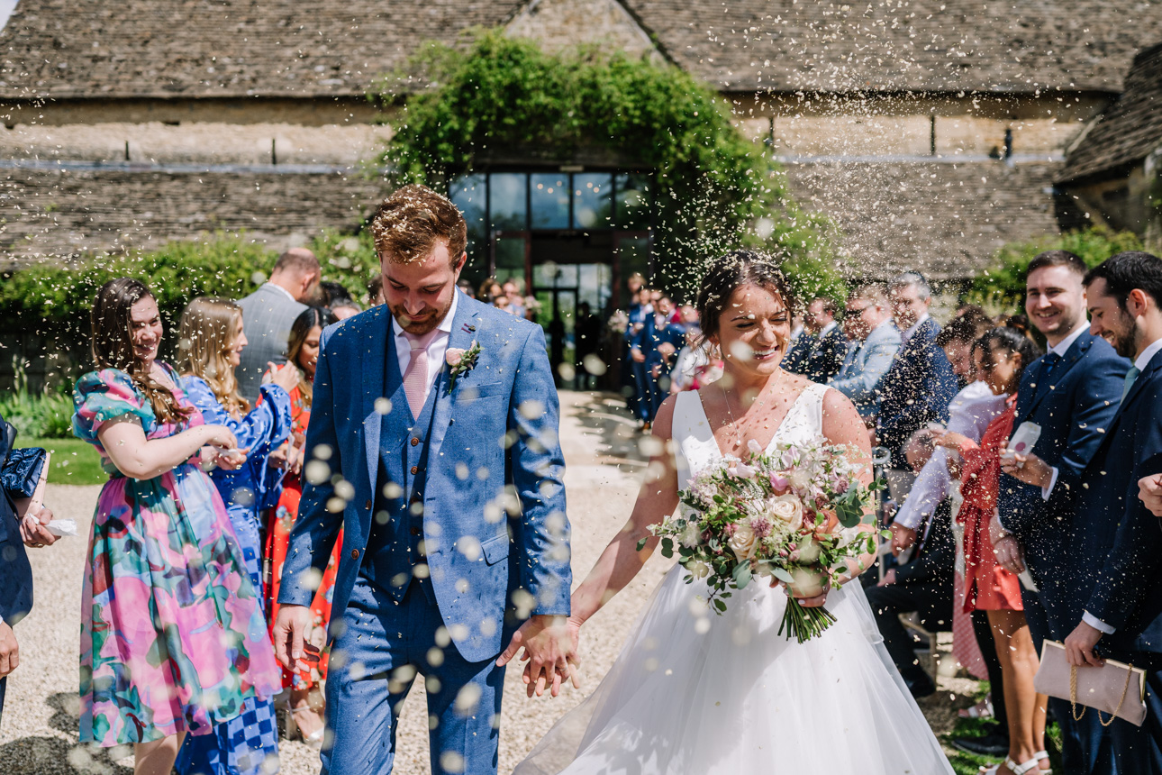 GREAT TYTHE BARN WEDDING PHOTOGRAPHER 31 Olivia and Perry laughing as their wedding guests throw a massive cloud of colorful flower petal confetti outside at The Great Tythe Barn