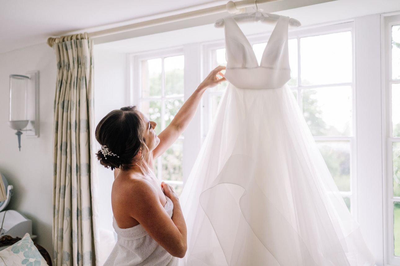 GREAT TYTHE BARN WEDDING PHOTOGRAPHER 19 A bride looking up at her wedding dress hanging elegantly against a large window during morning prep at The Great Tythe Barn accommodation.