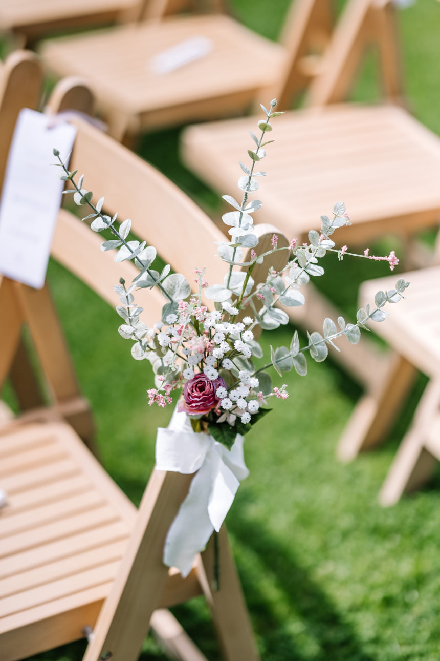 GREAT TYTHE BARN WEDDING PHOTOGRAPHER 36 Close-up of delicate floral arrangements tied with white ribbons onto wooden aisle chairs for an outdoor wedding ceremony at The Great Tythe Barn.
