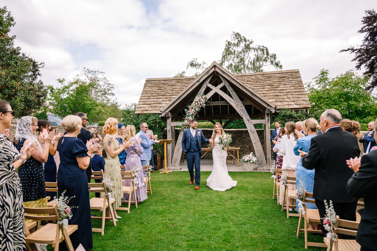 GREAT TYTHE BARN WEDDING PHOTOGRAPHER 42 Newlyweds Rachel and Leigh smiling and holding hands as they walk back down the aisle together after their outdoor ceremony at The Great Tythe Barn.