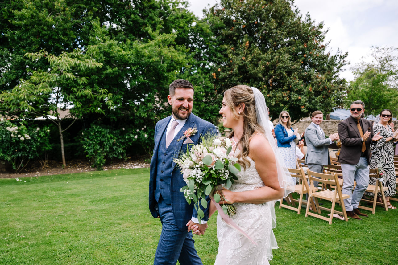 GREAT TYTHE BARN WEDDING PHOTOGRAPHER 43 Rachel and Leigh laughing together on the grass while surrounded by lush green trees during their reception at The Great Tythe Barn.