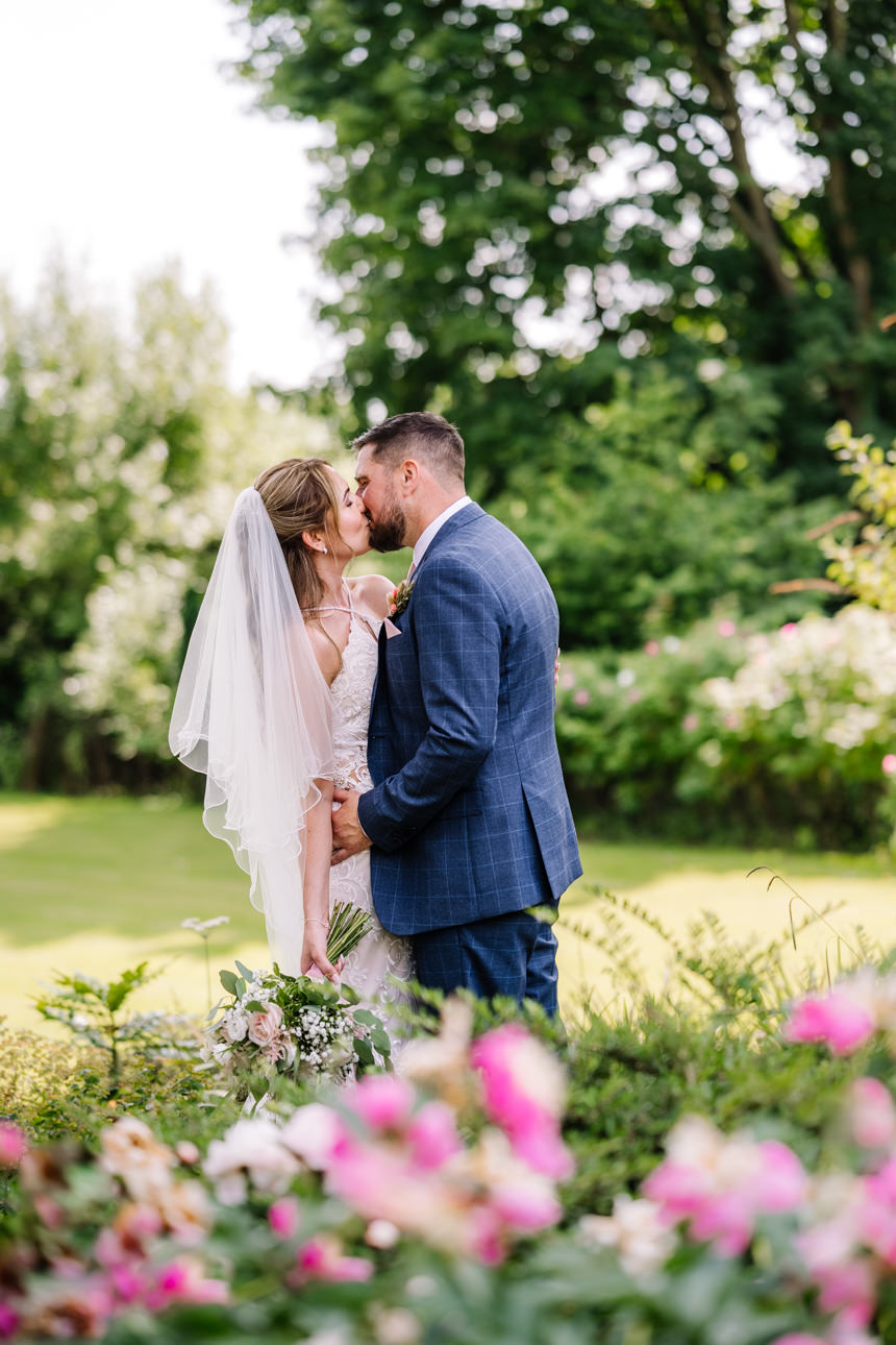 GREAT TYTHE BARN WEDDING PHOTOGRAPHER 69 The newlyweds walking hand-in-hand past lush green hedges and flowers during their summer wedding at The Great Tythe Barn. Photographed by Greta Tythe Barn Wedding Photographer, Tanli Joy Weddings.