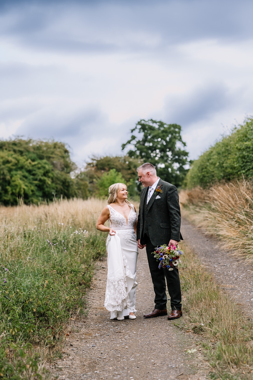 GREAT TYTHE BARN WEDDING PHOTOGRAPHER 5 A bride and groom walking down a rustic dirt pathway surrounded by summer fields on the grounds of The Great Tythe Barn.