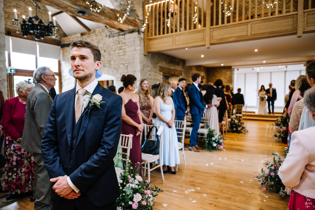 GREAT TYTHE BARN WEDDING PHOTOGRAPHER 23 A groom in a navy suit standing inside the barn at The Great Tythe Barn, waiting for his bride to arrive for the wedding ceremony.
