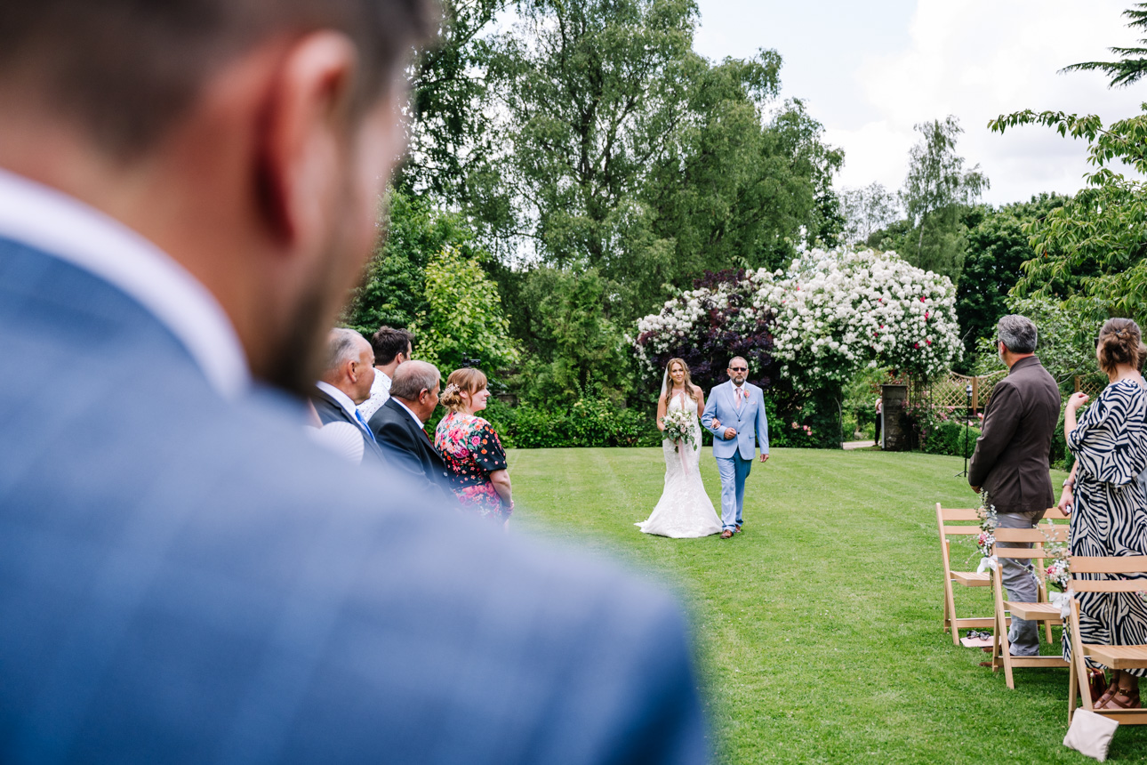 GREAT TYTHE BARN WEDDING PHOTOGRAPHER 41 An over-the-shoulder portrait of a groom looking down the aisle toward the outdoor pavilion setup at The Great Tythe Barn.
