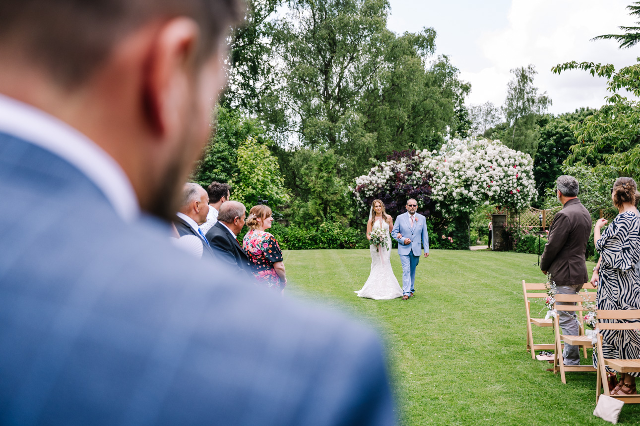GREAT TYTHE BARN WEDDING PHOTOGRAPHER 11 Over-the-shoulder portrait of a groom looking at his bride walking down the aisle at The Great Tythe Barn.