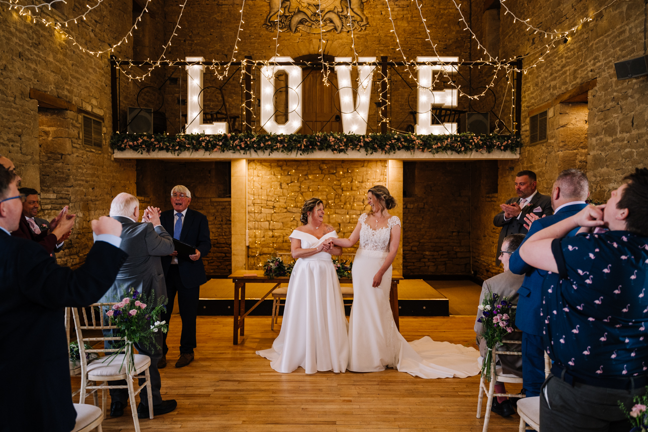 GREAT TYTHE BARN WEDDING PHOTOGRAPHER 32 Two brides dancing their romantic first dance in front of a giant illuminated LOVE sign beneath the fairy lights at The Great Tythe Barn.