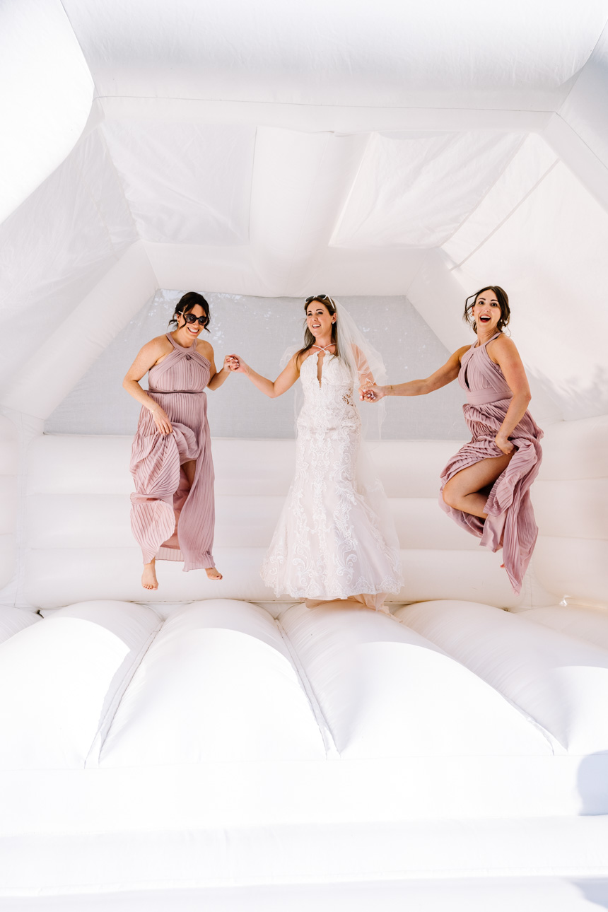 GREAT TYTHE BARN WEDDING PHOTOGRAPHER 101 A group of bridesmaids in soft pink dresses joyfully laughing and bouncing on a bouncy castle at the Great Tythe Barn in tetbury.