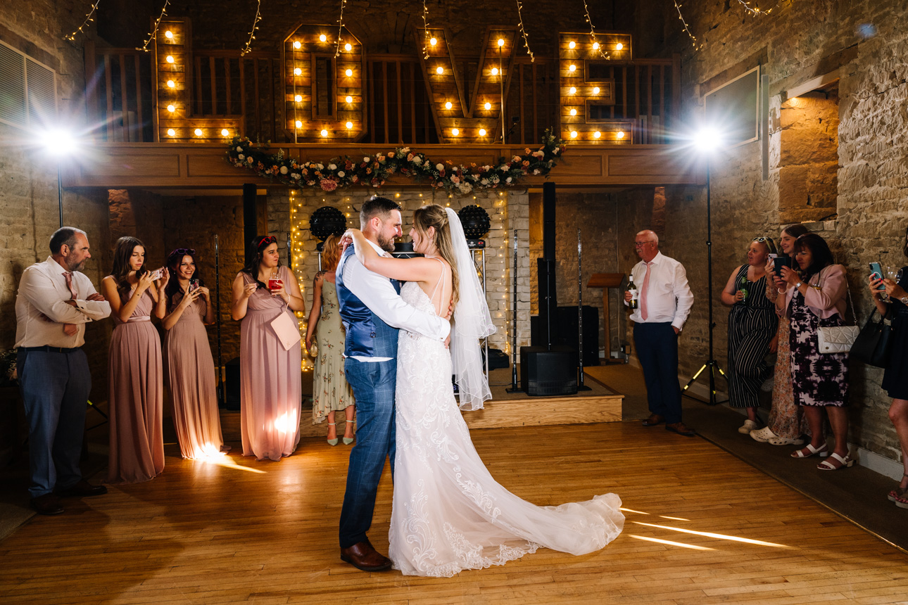 GREAT TYTHE BARN WEDDING PHOTOGRAPHER 72 A bride and groom sharing their romantic first dance beneath a canopy of fairy lights and an illuminated LOVE sign inside The Great Tythe Barn.