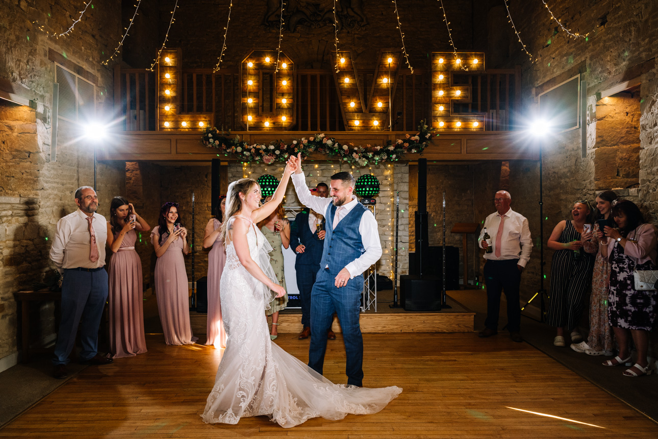 GREAT TYTHE BARN WEDDING PHOTOGRAPHER 4 A bride and groom sharing their romantic first dance inside the great tythe barn beneath a massive canopy of warm, twinkling fairy lights and a "LOVE" light-up sign.